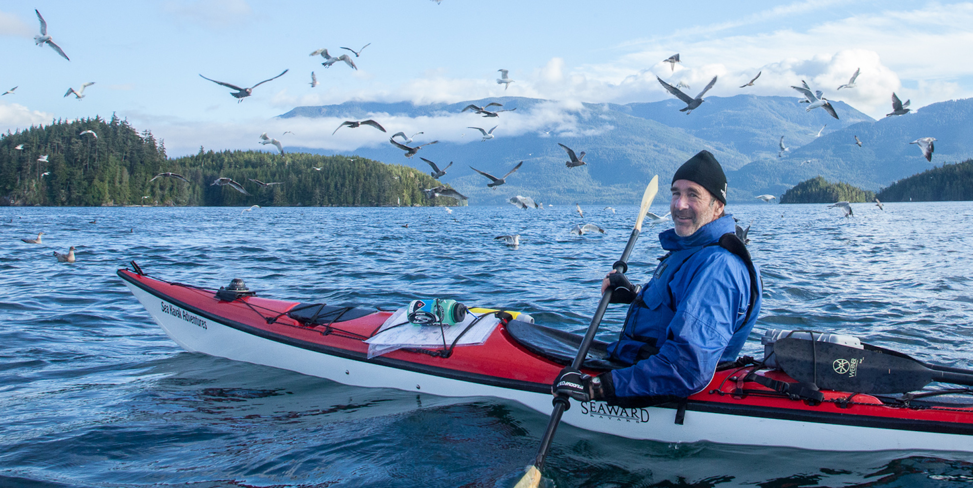 A man in a single red kayak with a blue dry top and black beanie smiling for the camera while a herd of birds fly overhead