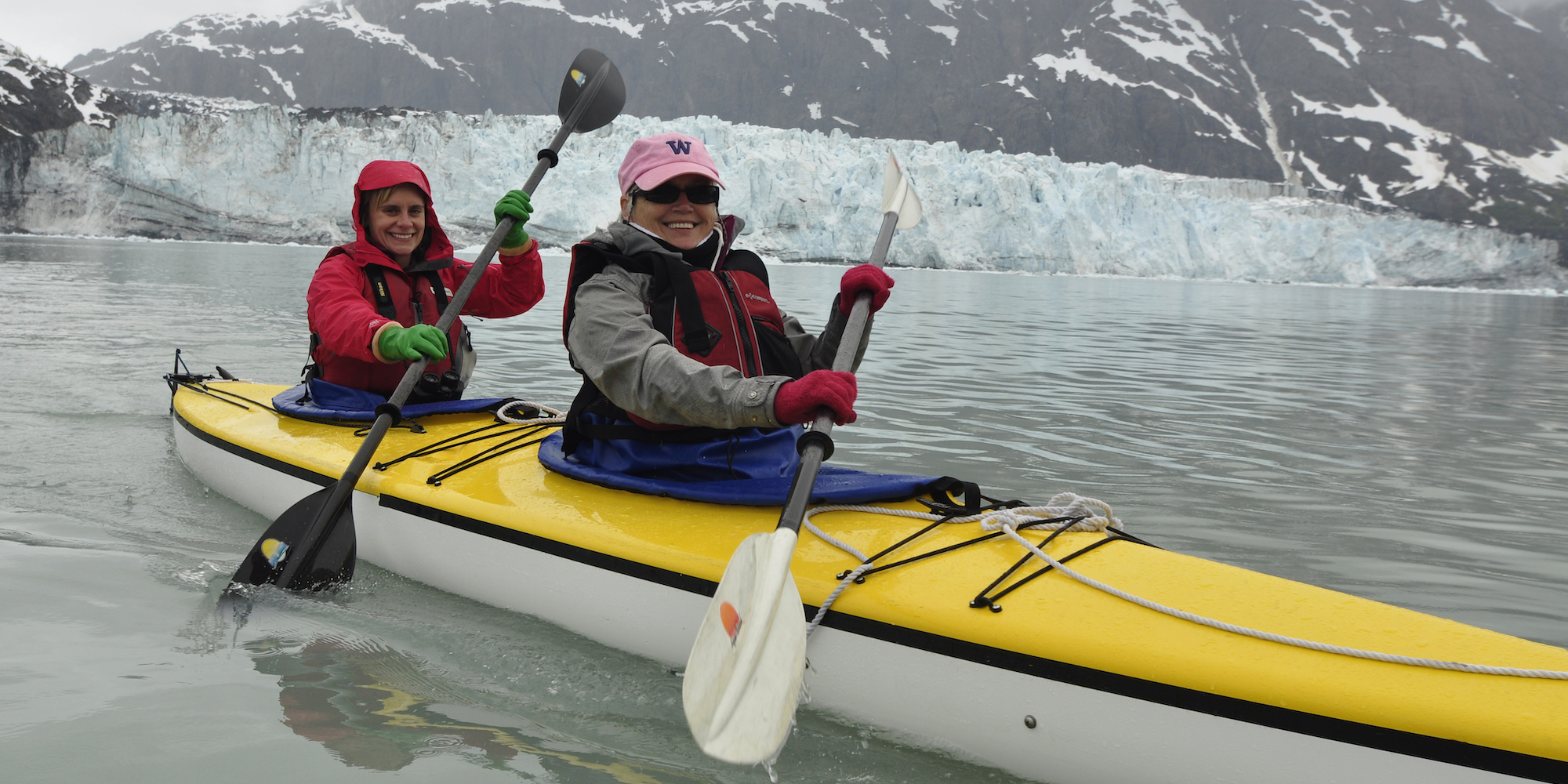 Two women paddling a yellow tandem kayak both wearing colorful gloves in Alaska in front of glaciers