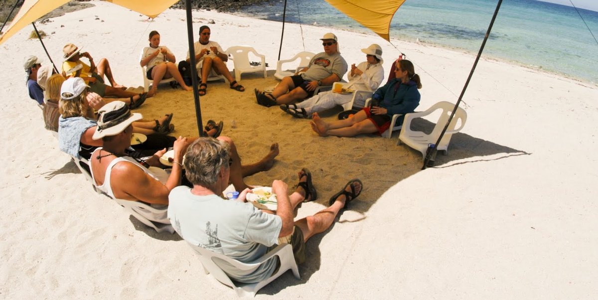 A group of sea kayakers sitting in the sand in a circle under a shade tarp