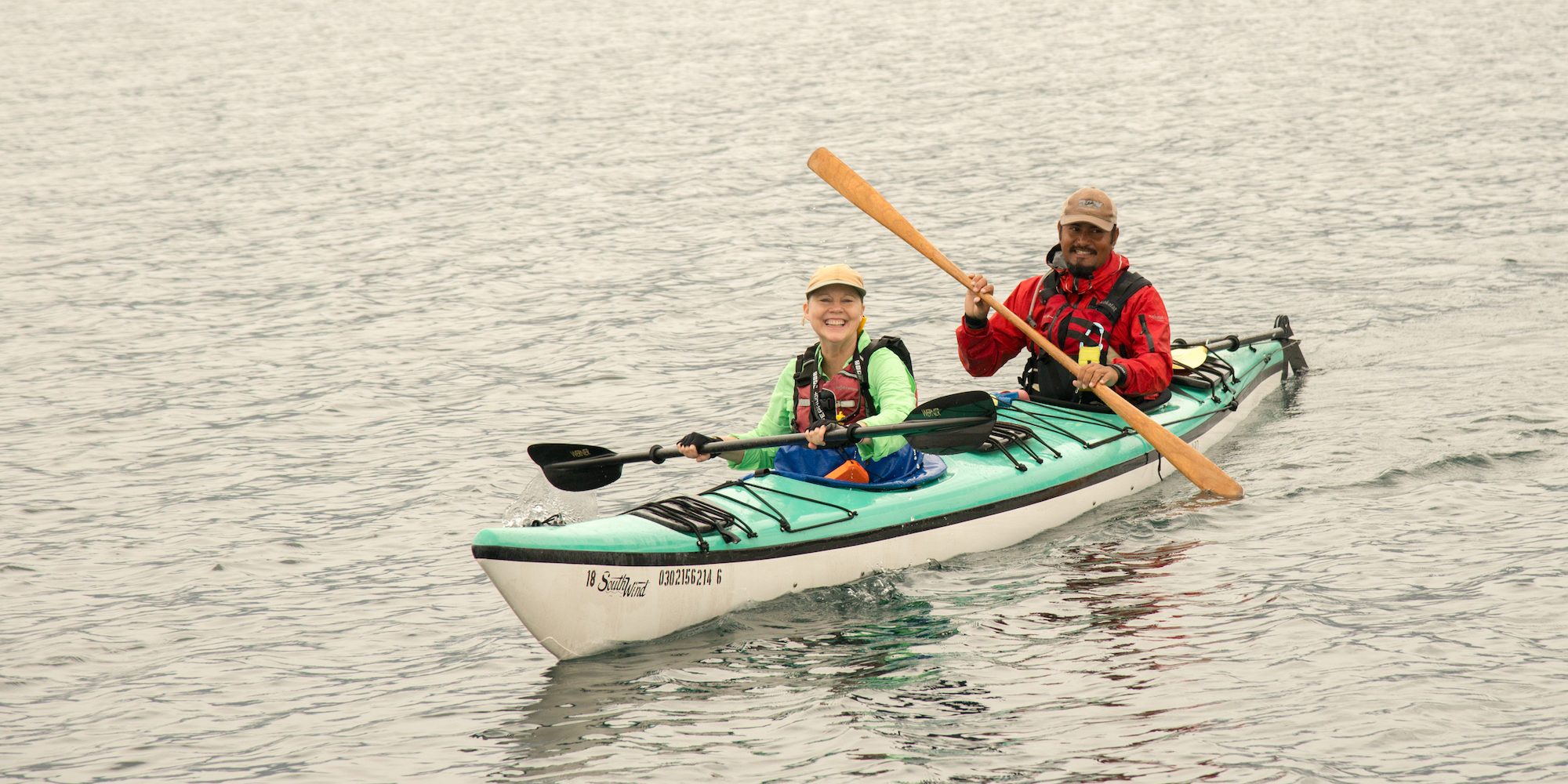 Two people in a tandem kayak smiling while holding different paddles, one wooden and one plastic.