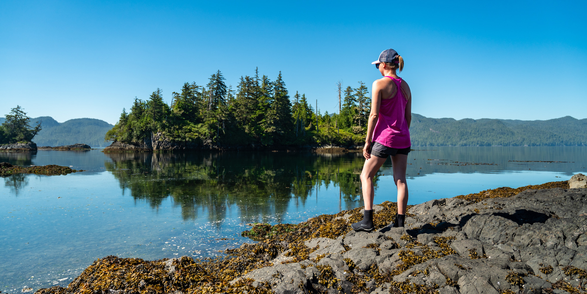 A woman standing along a rocky shore while on a hike on a sunny day in God's Pocket Provincial Park in British Columbia
