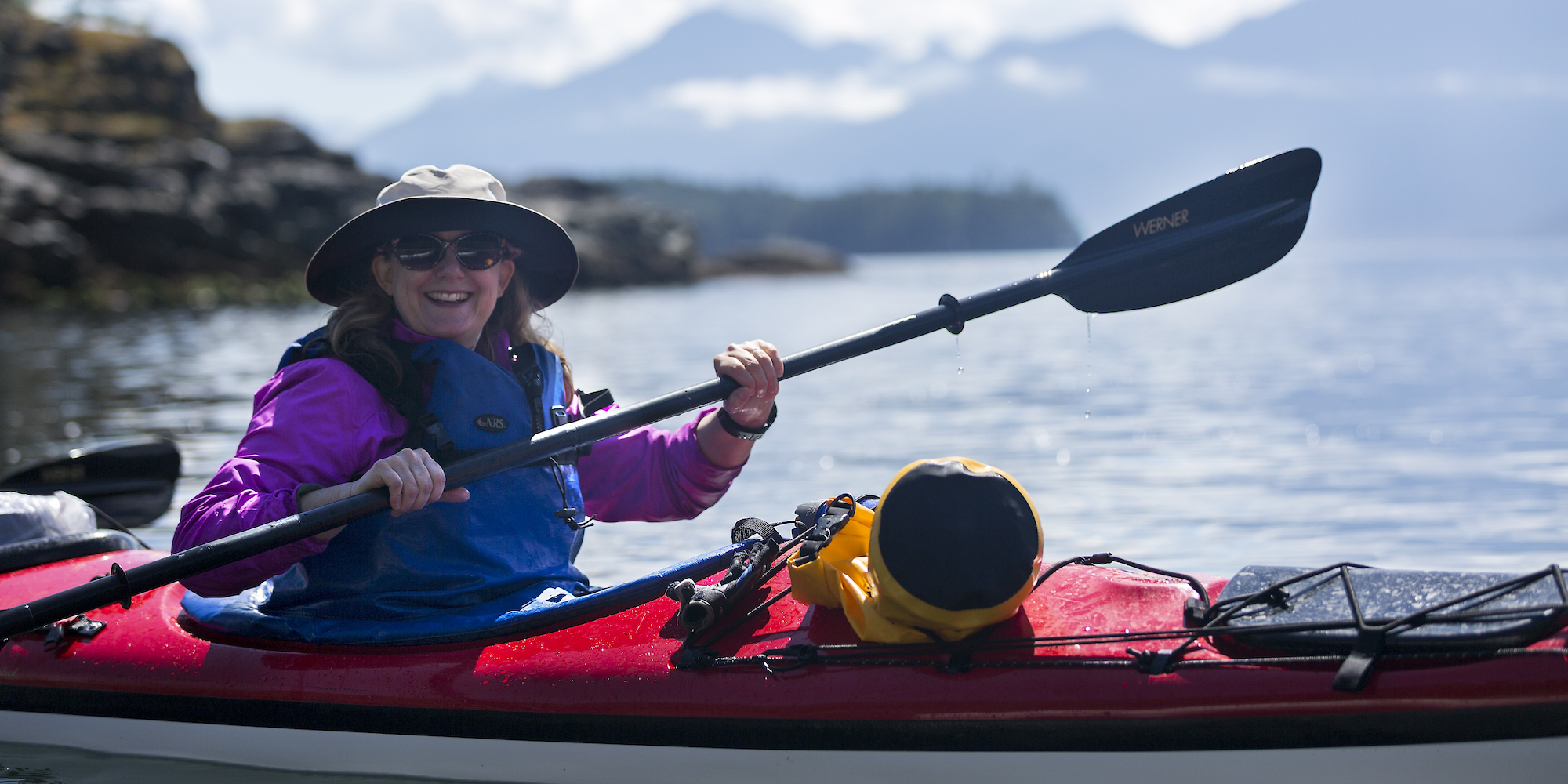 A women in a red sea kayak on a sunny day holding up her paddle and smiling