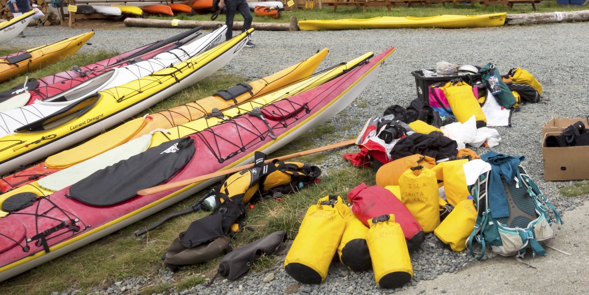A row of yellow, white, and red sea kayaking pulled up onto a rocky shore with a group of small yellow dry bags next to them on shore