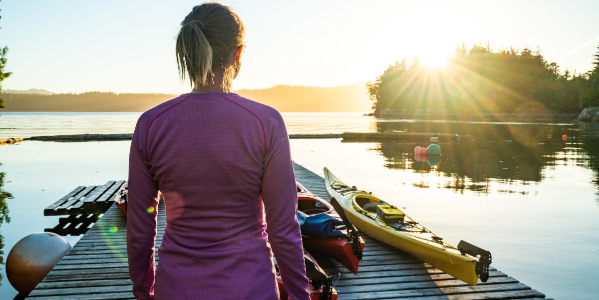 Girl from behind standing on a dock in a pink synthetic shirt watching the sunrise over the kayaks tied to the dock
