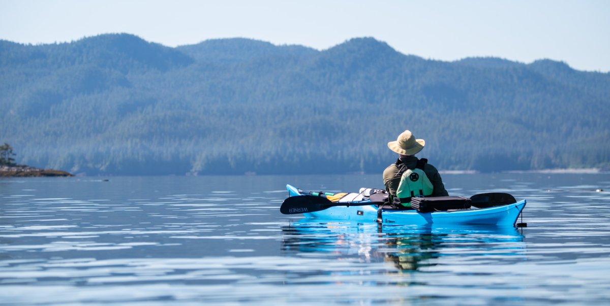 Picture of a person sea kayaking from behind wearing a sun hat overlooking forested hills in British Columbia