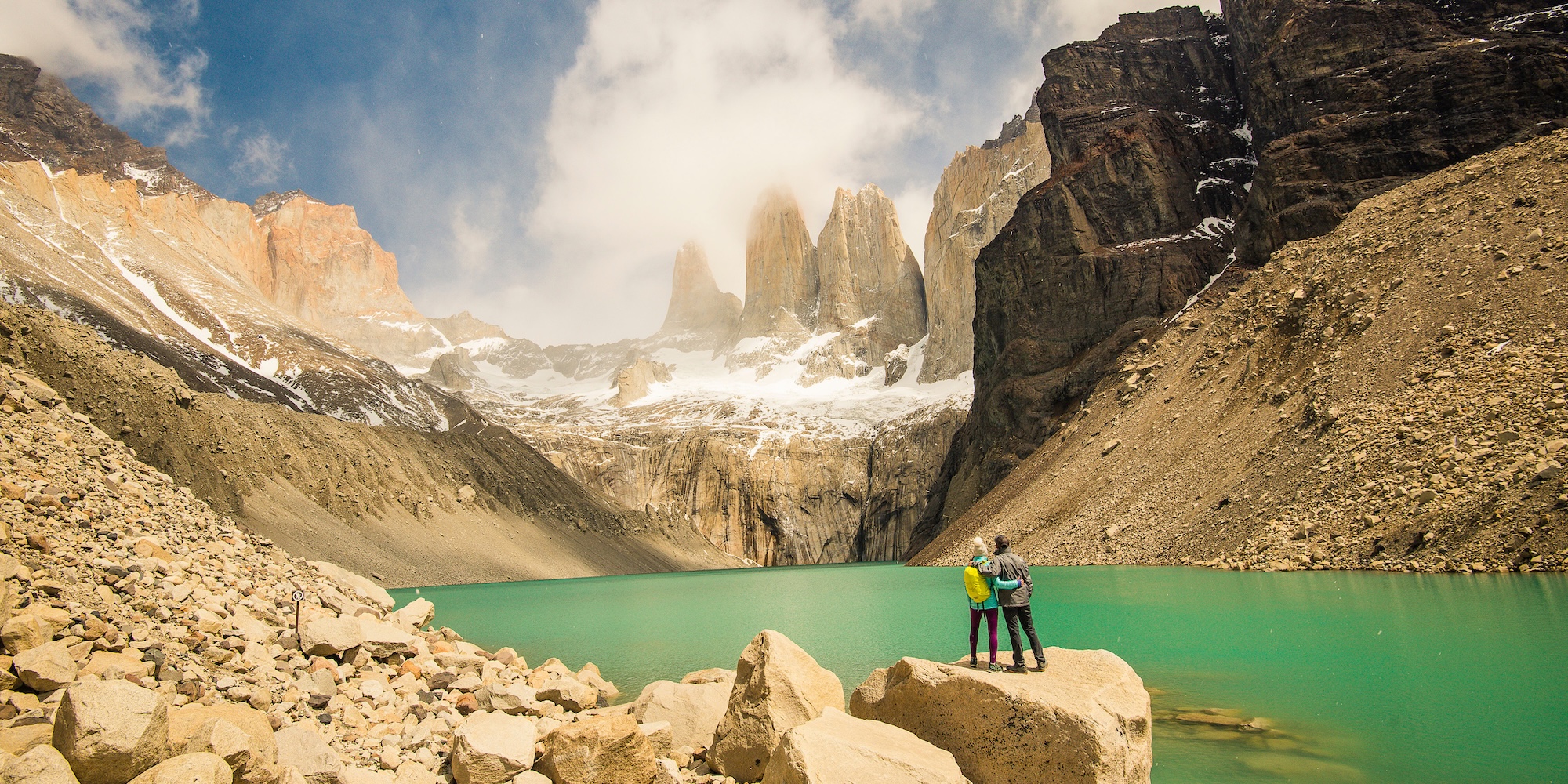 Two hikers standing on rocks at the base of the iconic Las Torres peaks, with a turquoise glacial lake below—one of the top trekking highlights in Patagonia.