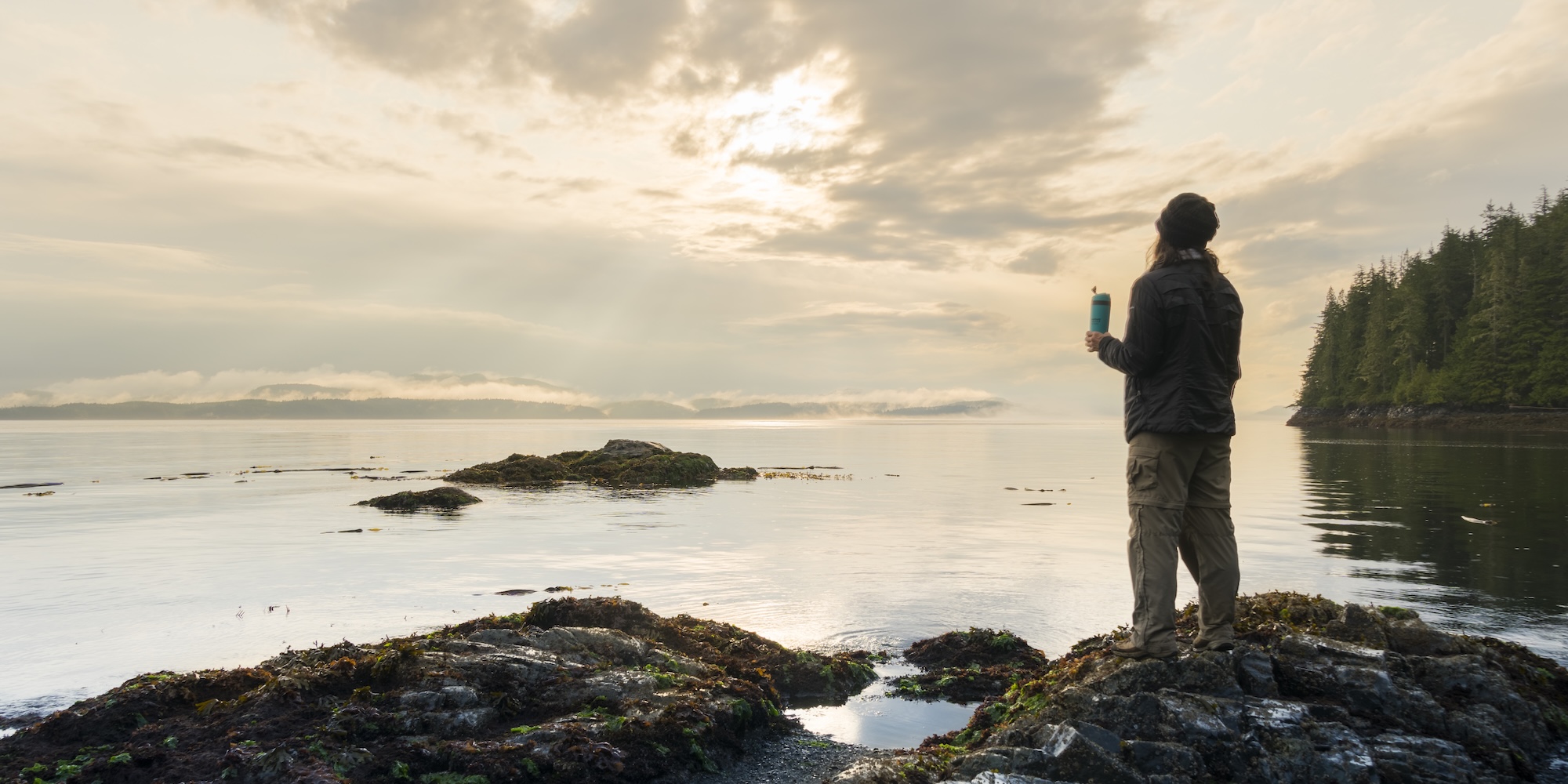 Person standing on a rocky shoreline at sunrise overlooking calm ocean waters on Vancouver Island, British Columbia