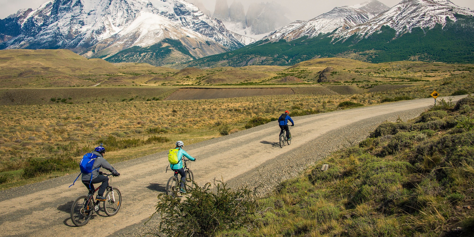 Three cyclists riding along a gravel road in Patagonia with the Andes in the distance, showcasing the type of active outdoor gear ideal for exploring the region.