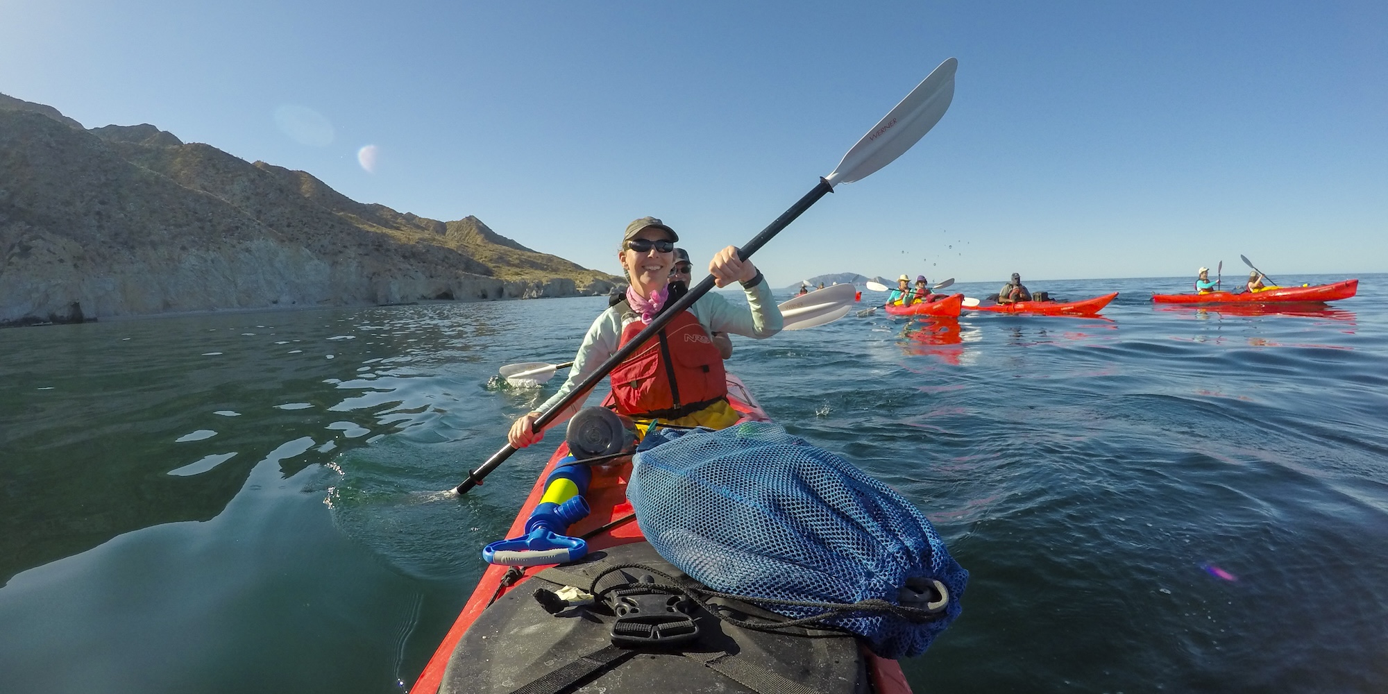 Two people paddle a red tandem kayak on calm blue water near rocky cliffs, smiling under a clear sky. Other kayakers follow in the distance, all wearing life jackets.