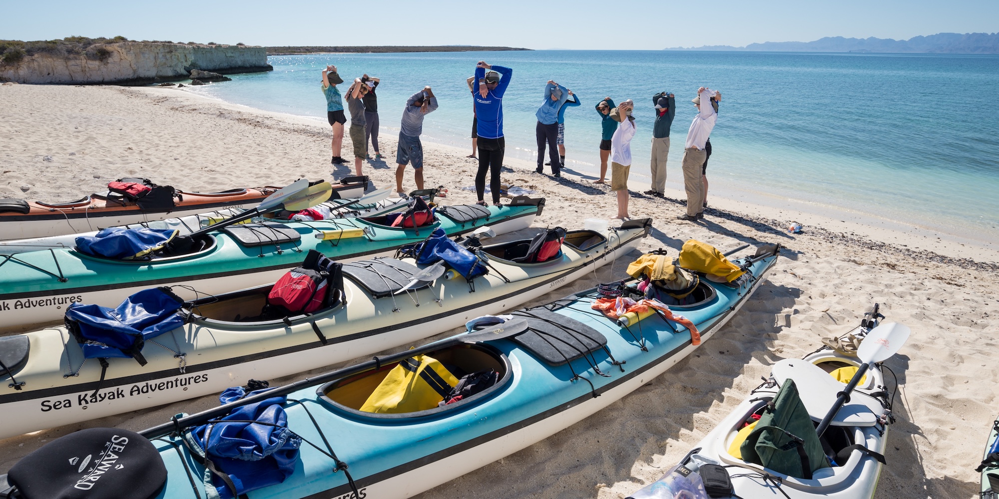 A group of sea kayakers on a sandy beach in Baja stretching before a day of paddling.