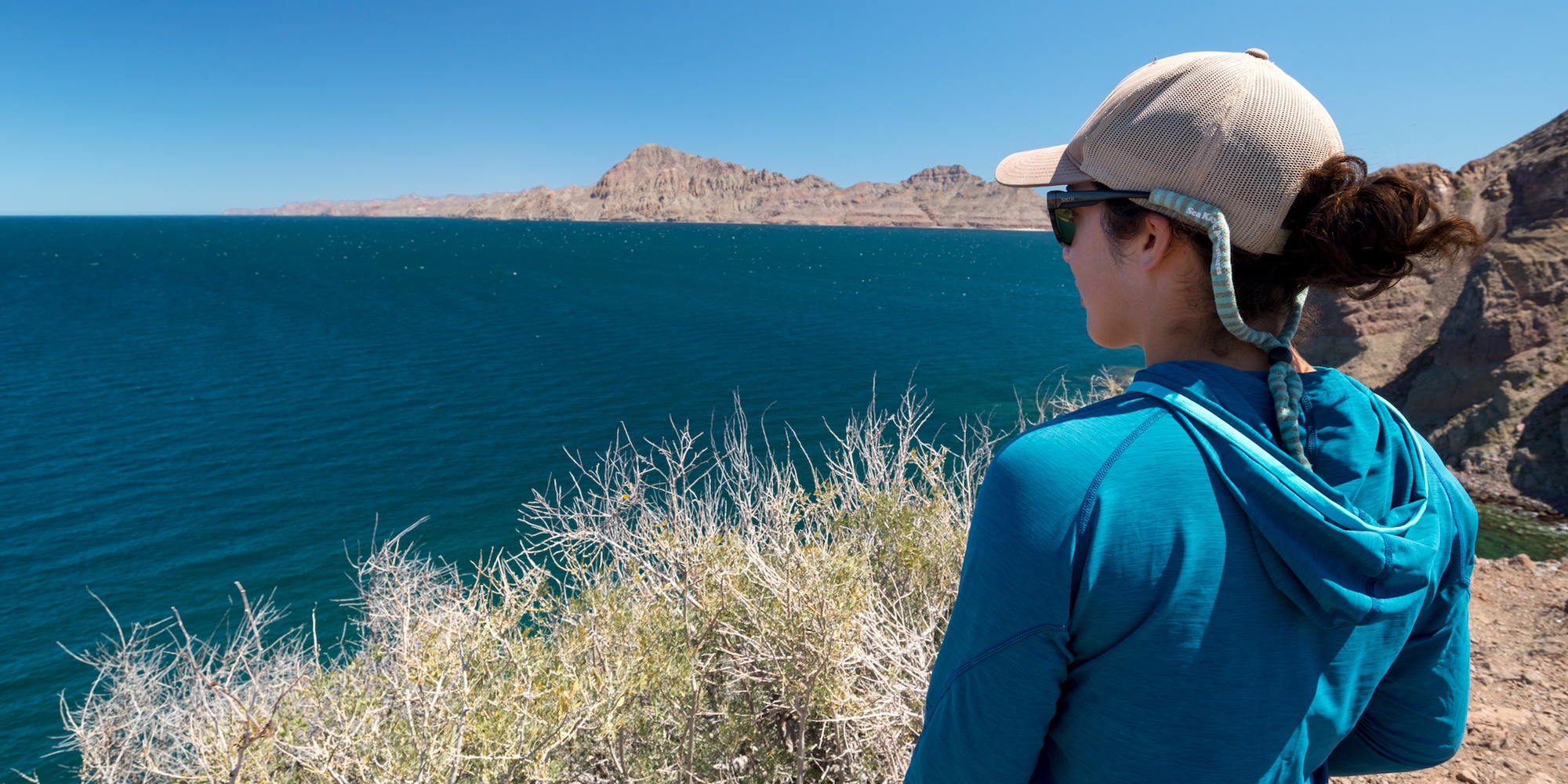 Female traveler in a blue sun shirt and cap looking out over the Sea of Cortez, with rugged desert cliffs and deep blue water stretching into the distance.