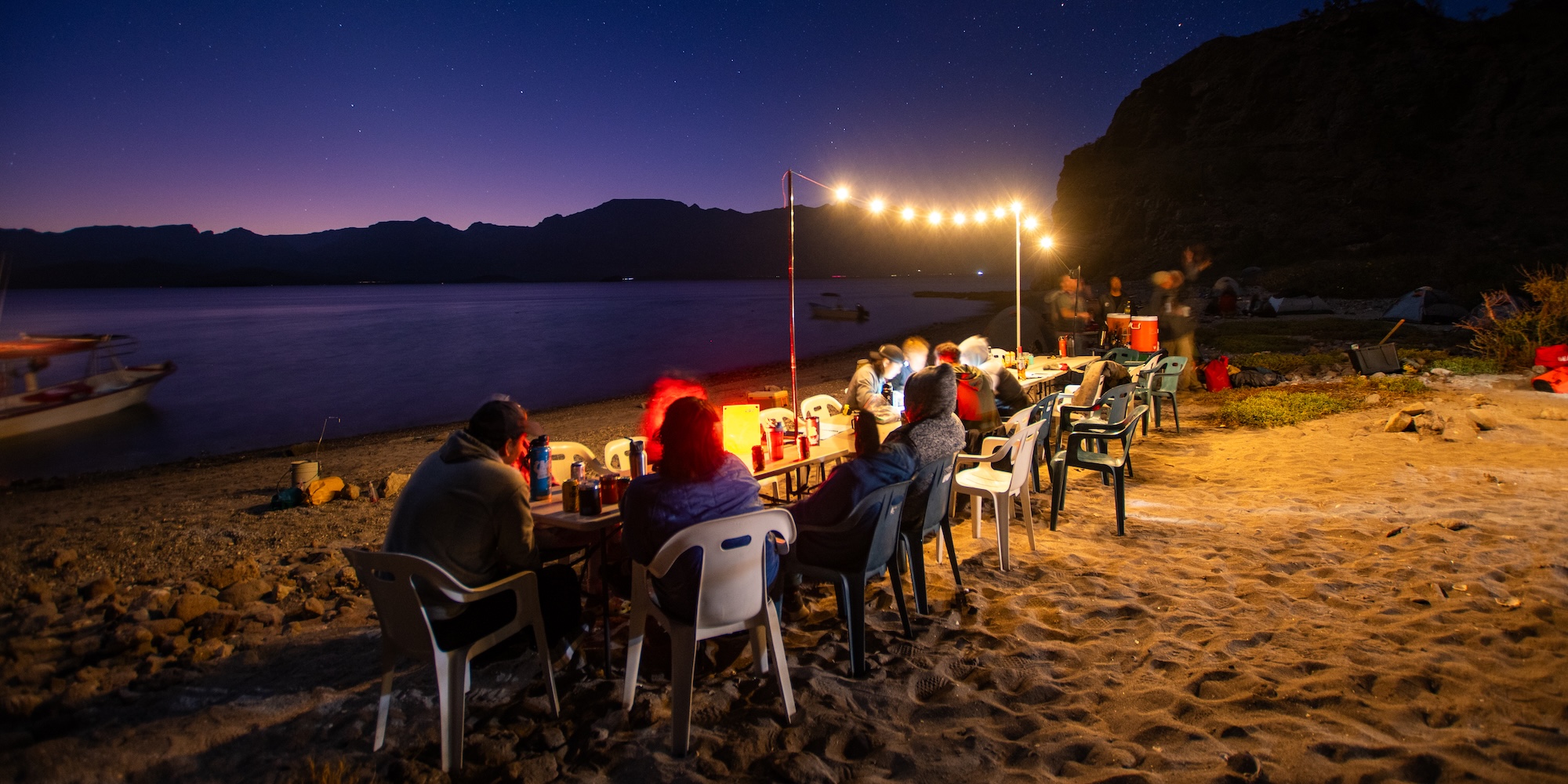 Guests enjoy a communal dinner under string lights on a sandy beach at dusk, with calm water and distant mountains silhouetted against a starry sky.