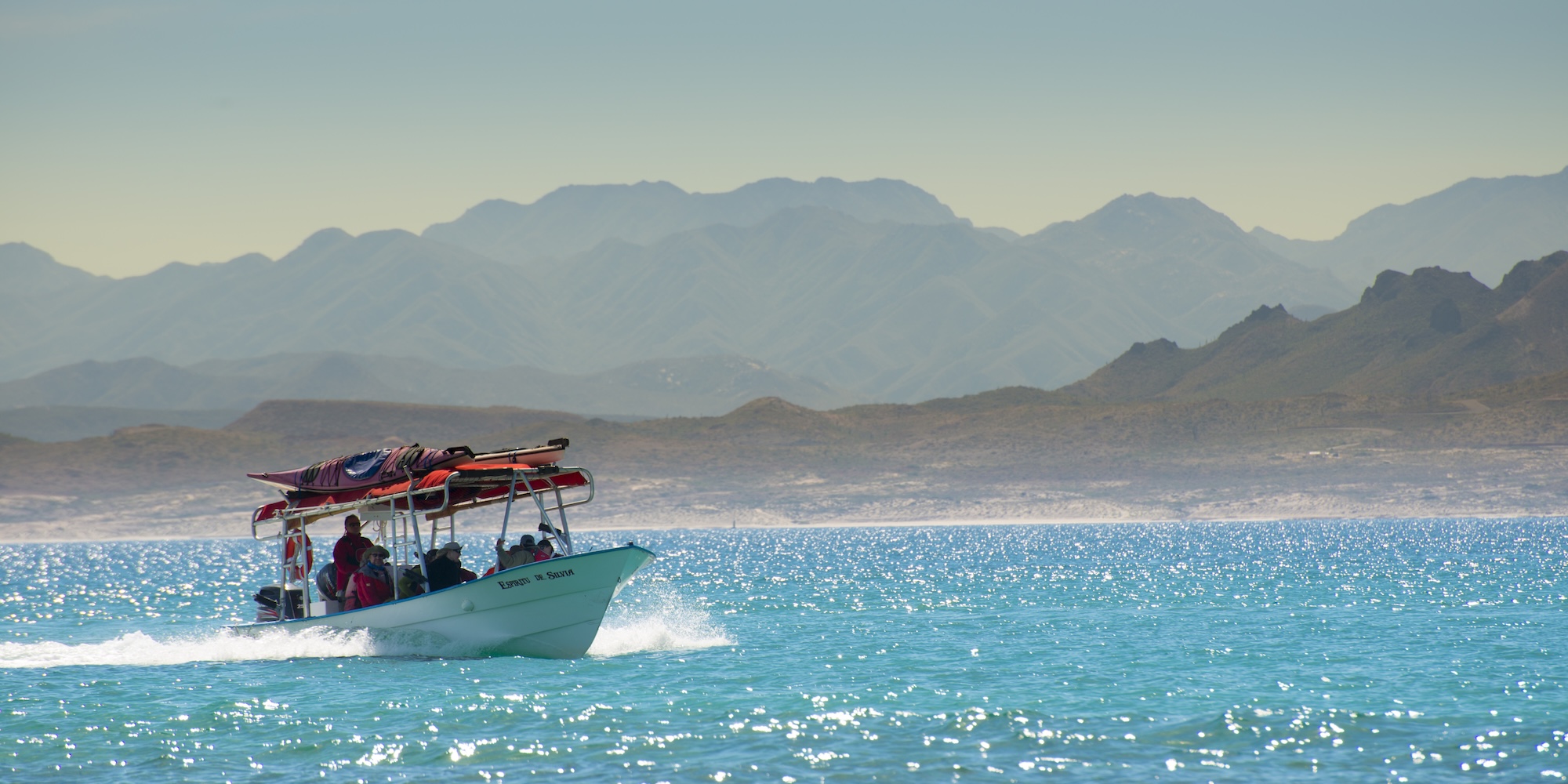 A panga support boat carries kayakers across bright turquoise water with mountainous desert terrain in the background. The sun glistens on the sea as the boat moves at speed.