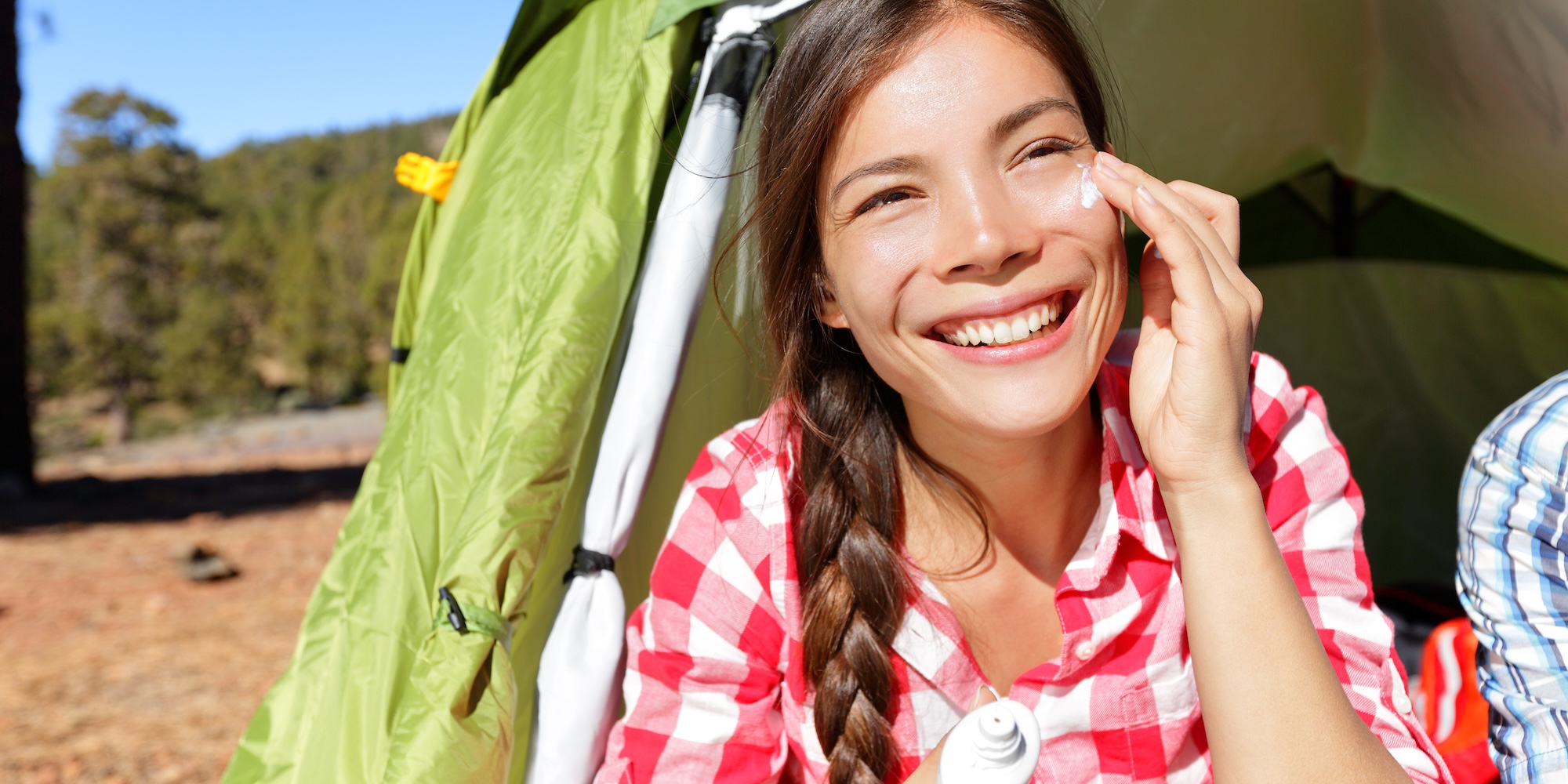 Smiling woman sitting outside a tent, applying sunscreen to her face during a sunny day of camping.