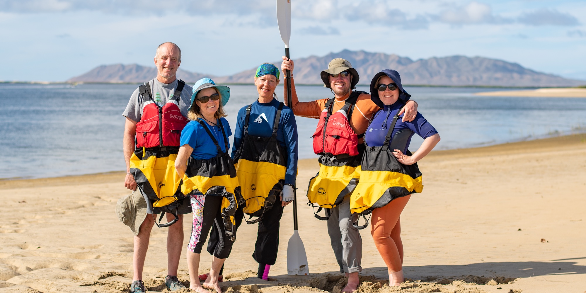 A smiling group of five kayakers poses together on a sunny beach, dressed in splash gear with a paddle held upright, with calm ocean waters and distant hills in the background.