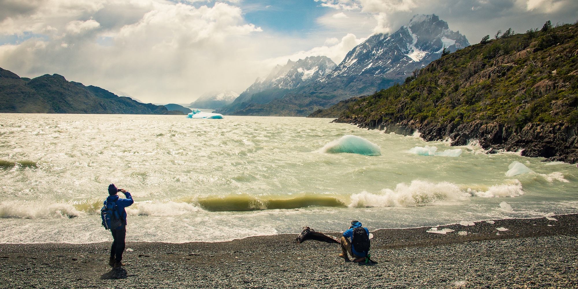 Two travelers on the windy shoreline of a glacial lake with floating icebergs and towering mountains in the background, illustrating Patagonia’s unpredictable weather.