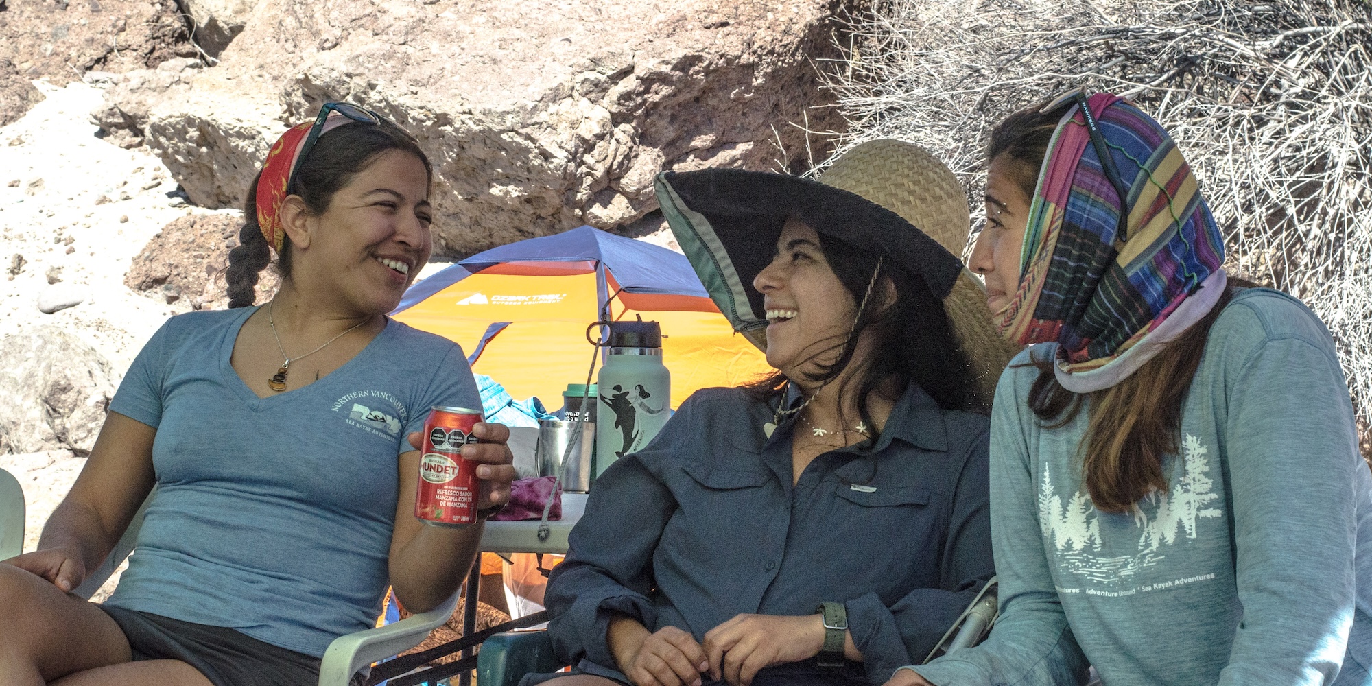 Three women sharing a laugh while relaxing at a campsite in Baja, with colorful tents and desert rock formations in the background.