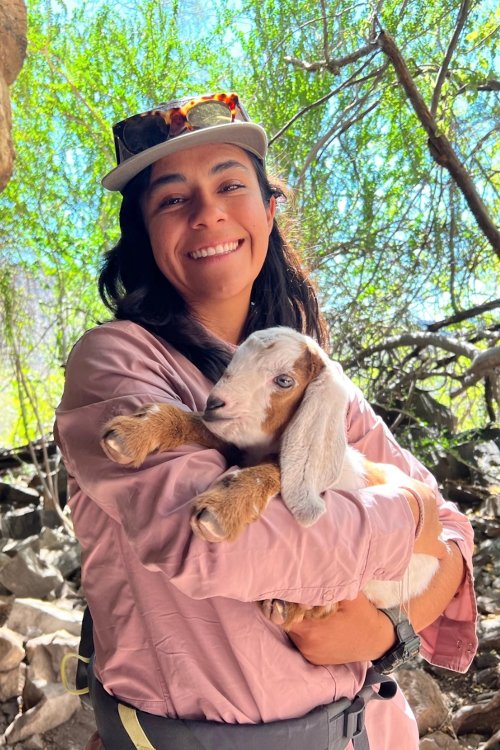 Guide holding a baby goat outdoors in a shaded forest setting, smiling while surrounded by green trees and natural rocky terrain.