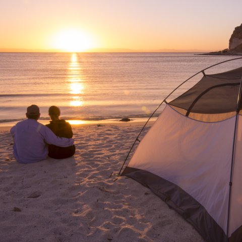 A couple sitting on a beach in front of a tent watching the sunset