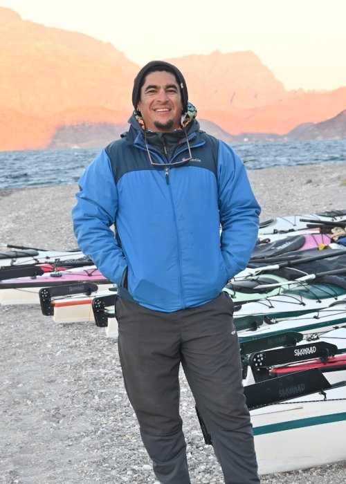 Baja Sea Kayak Guide standing on a beach in Loreto with sea kayaks lined up behind him. 