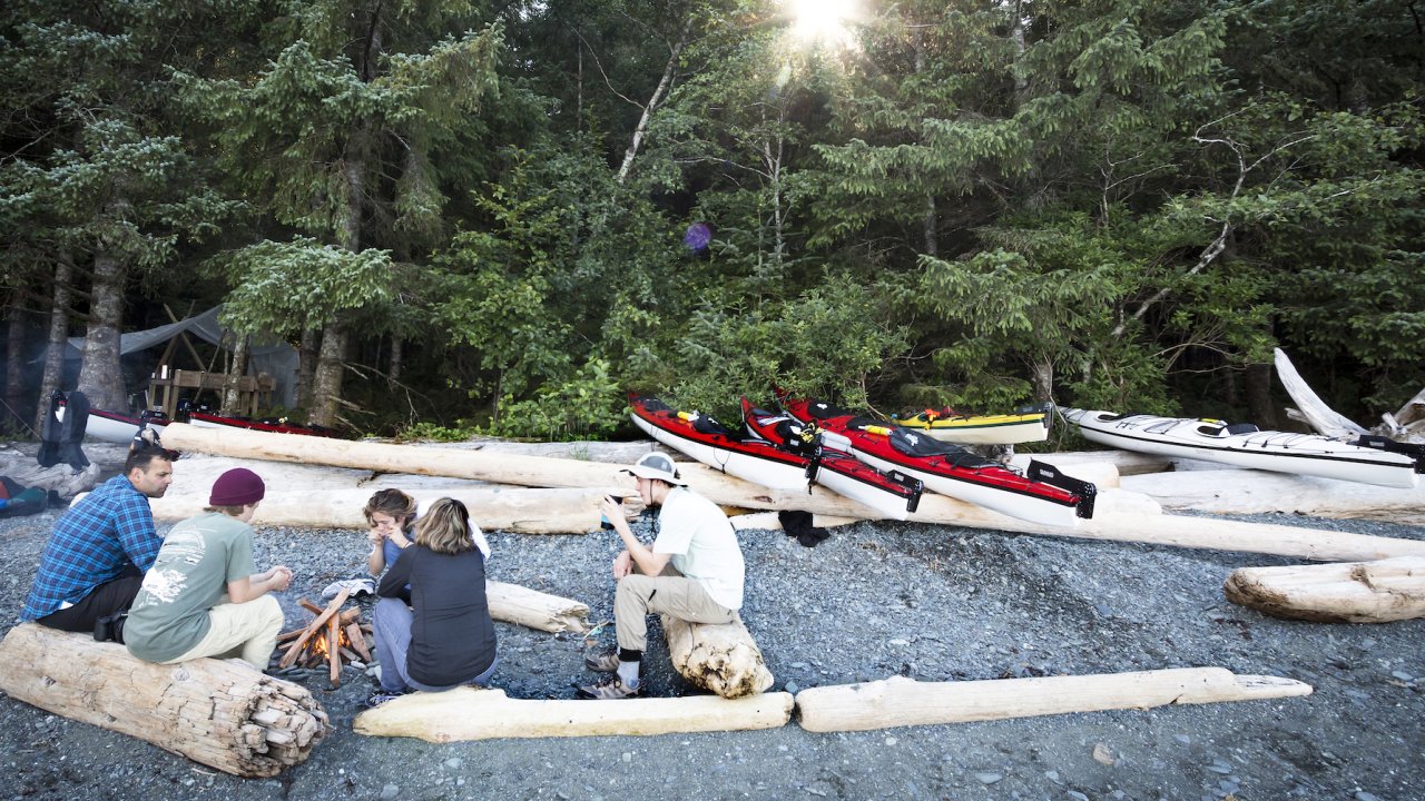A group of people sitting around a campfire on a rocky beach with their sea kayaks pulled on shore above logs