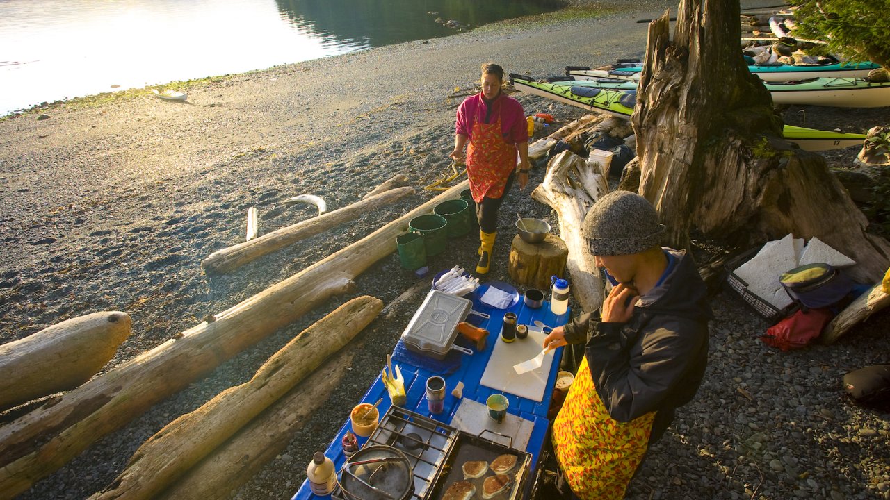 A cook partner stove on top of blue tables on a rocky beach while sea kayak guides cook at sunset