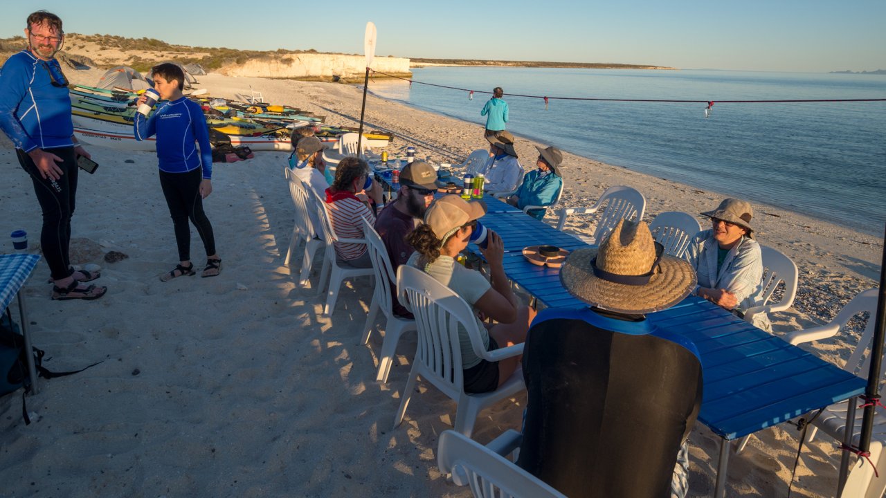 A group of people around a blue family style tent while camping on a beach on a sunny day