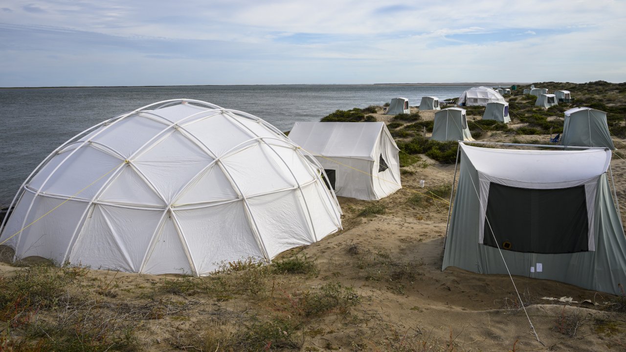 Dome style tent among other square canvas tents on a sand dune with the Pacific Ocean in the background