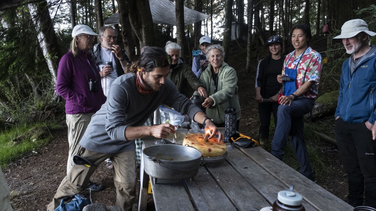 Guests gather around a picnic table at forest camp while a guide lights candles on a cake.