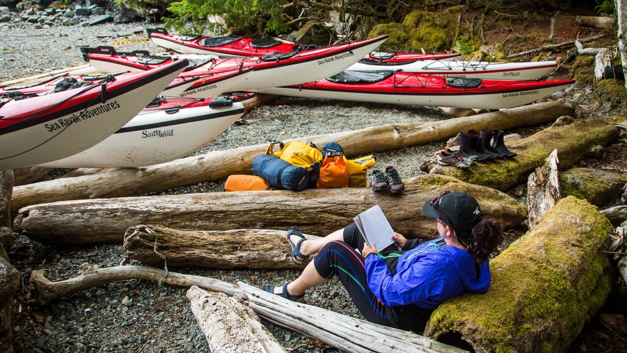 Guest reads on driftwood beside kayaks and dry bags at a British Columbia beach camp.