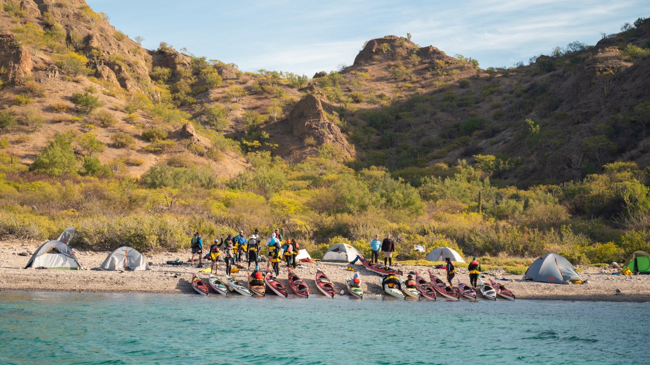 View of sea kayaks on shore set in front of a group of paddlers and tents in the Islands of Loreto Bay National Park