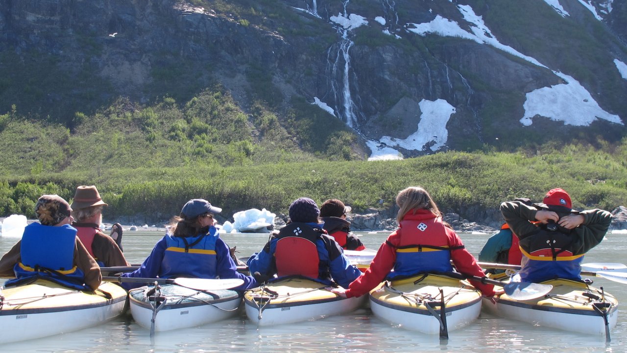 Group of people in yellow kayaks looking towards waterfalls, trees, and nearby mountains in Alaska
