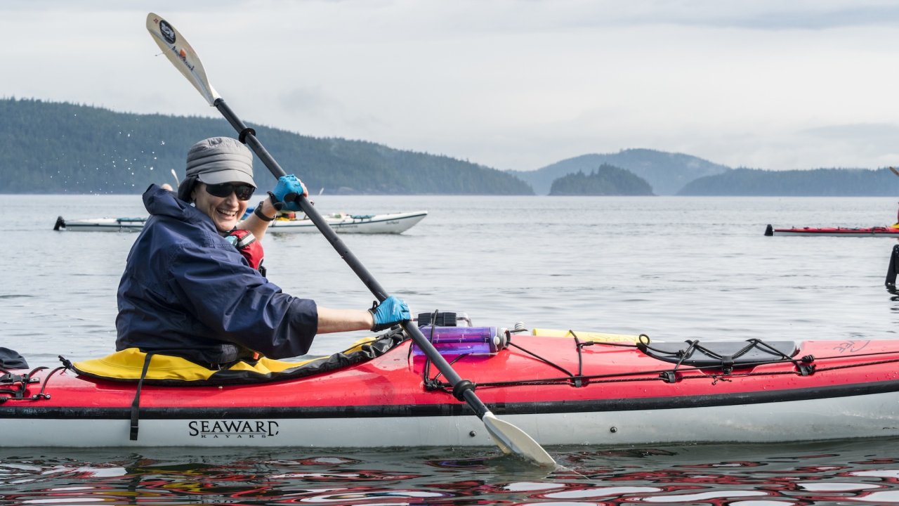 A smiling paddler in a red kayak enjoying scenic waters surrounded by forested islands, showcasing the adventure of kayaking Canada and British Columbia kayak tours.