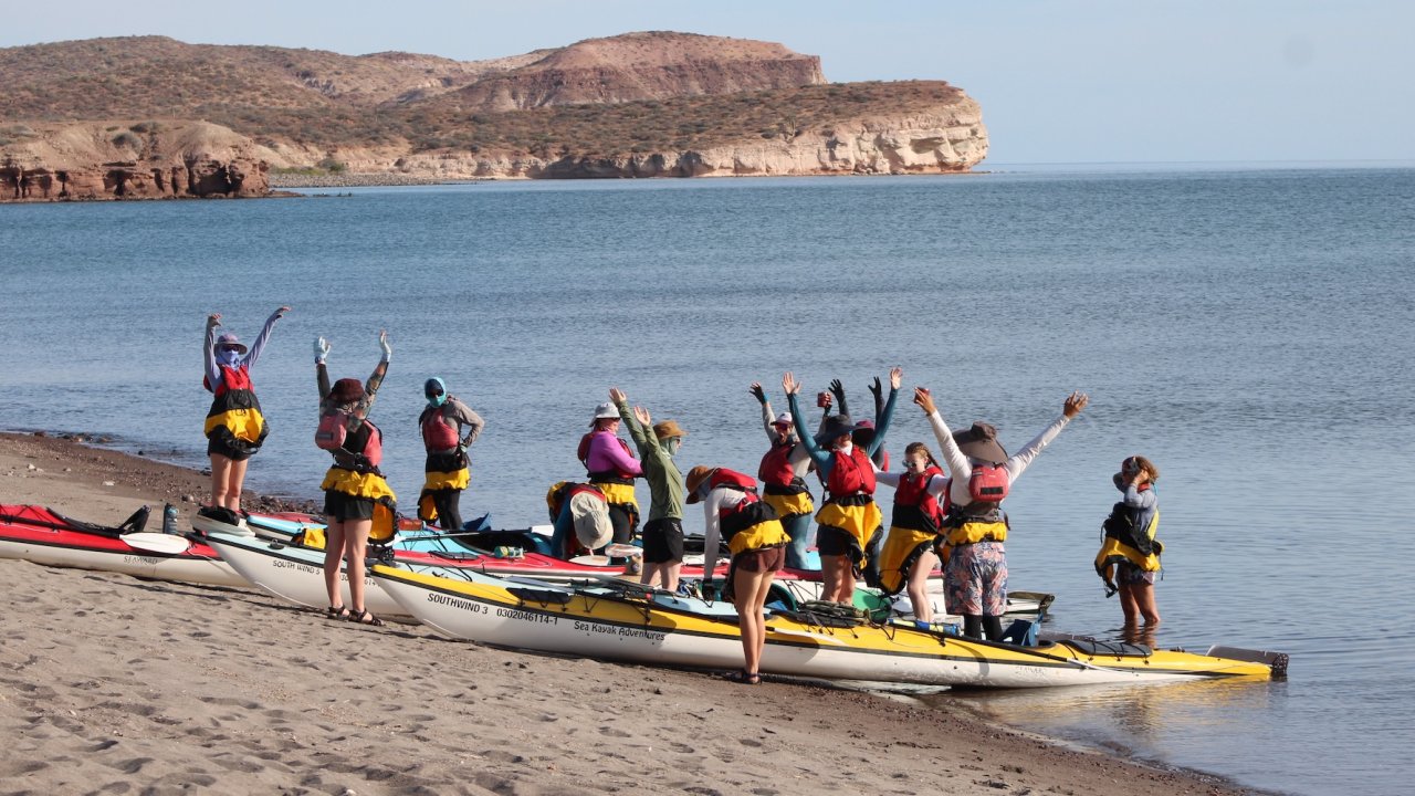 Group of student travelers standing beside sea kayaks on a sandy beach in Baja California Sur, preparing for a kayaking excursion along a calm coastline with rocky desert cliffs in the background.