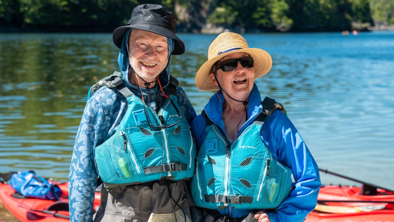 Two smiling kayakers in bright blue life jackets standing by red sea kayaks on the shore, ready to enjoy guided sea kayaking tours in British Columbia, Canada.
