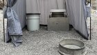  Metal burn pan in the sand next to two toilets inside of a stand up tent