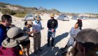 Group of people on the beach standing in a circle in front of a row of tents on a sunny day