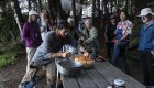 Guests gather around a picnic table at forest camp while a guide lights candles on a cake.