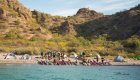 View of sea kayaks on shore set in front of a group of paddlers and tents in the Islands of Loreto Bay National Park