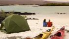 A couple sitting on a white sand beach with their tent and two sea kayaks behind them
