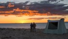 A couple standing next to a canvas tent watching the sunset over the Pacific Ocean