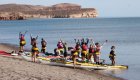 Group of student travelers standing beside sea kayaks on a sandy beach in Baja California Sur, preparing for a kayaking excursion along a calm coastline with rocky desert cliffs in the background.