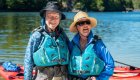 Two smiling kayakers in bright blue life jackets standing by red sea kayaks on the shore, ready to enjoy guided sea kayaking tours in British Columbia, Canada.