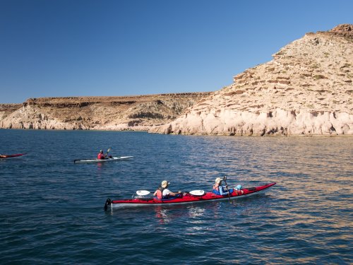 Sea kayakers paddle tandem and single kayaks across calm blue water in Baja’s Gulf of California, with desert cliffs rising along the shoreline during a sea kayaking trip.